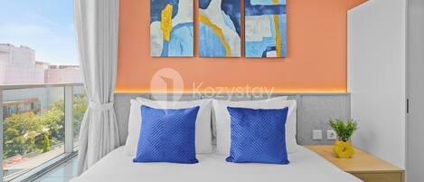 Quarto de hotel moderno com uma cama grande, duas almofadas azuis, uma mesa de cabeceira com uma planta e arte abstrata na parede.
