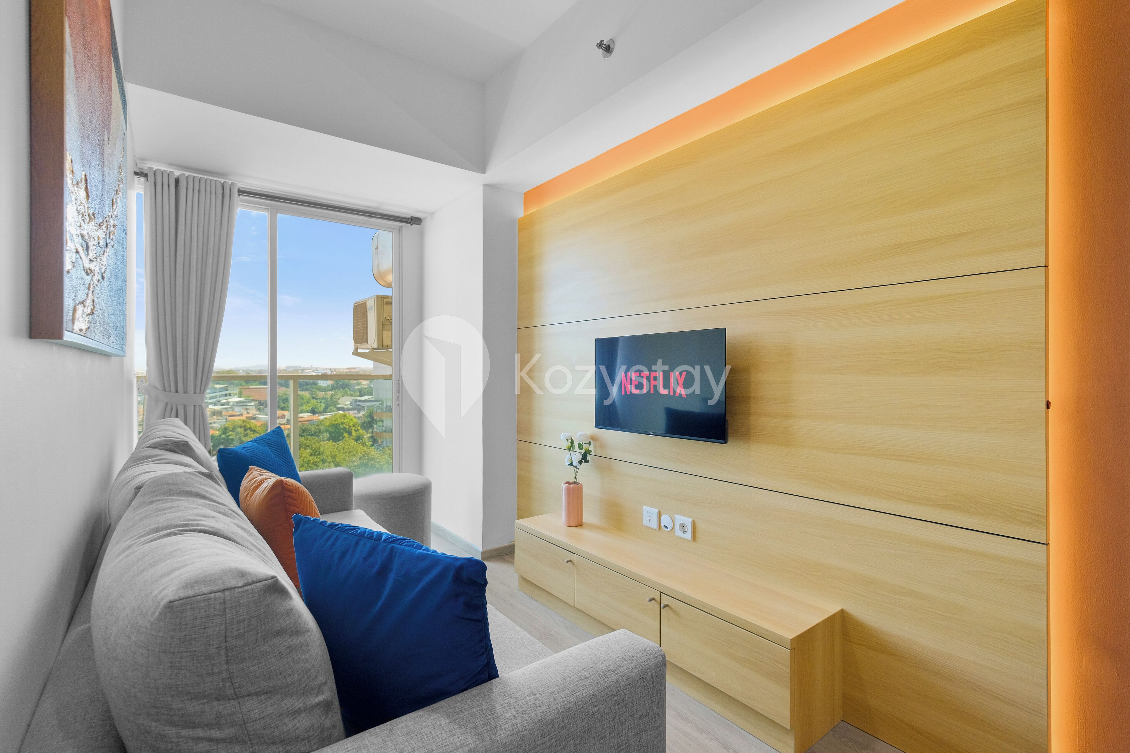 A modern living room with a grey sofa, blue cushions, a flat-screen TV on a wooden wall, and a view of a cityscape through large windows.