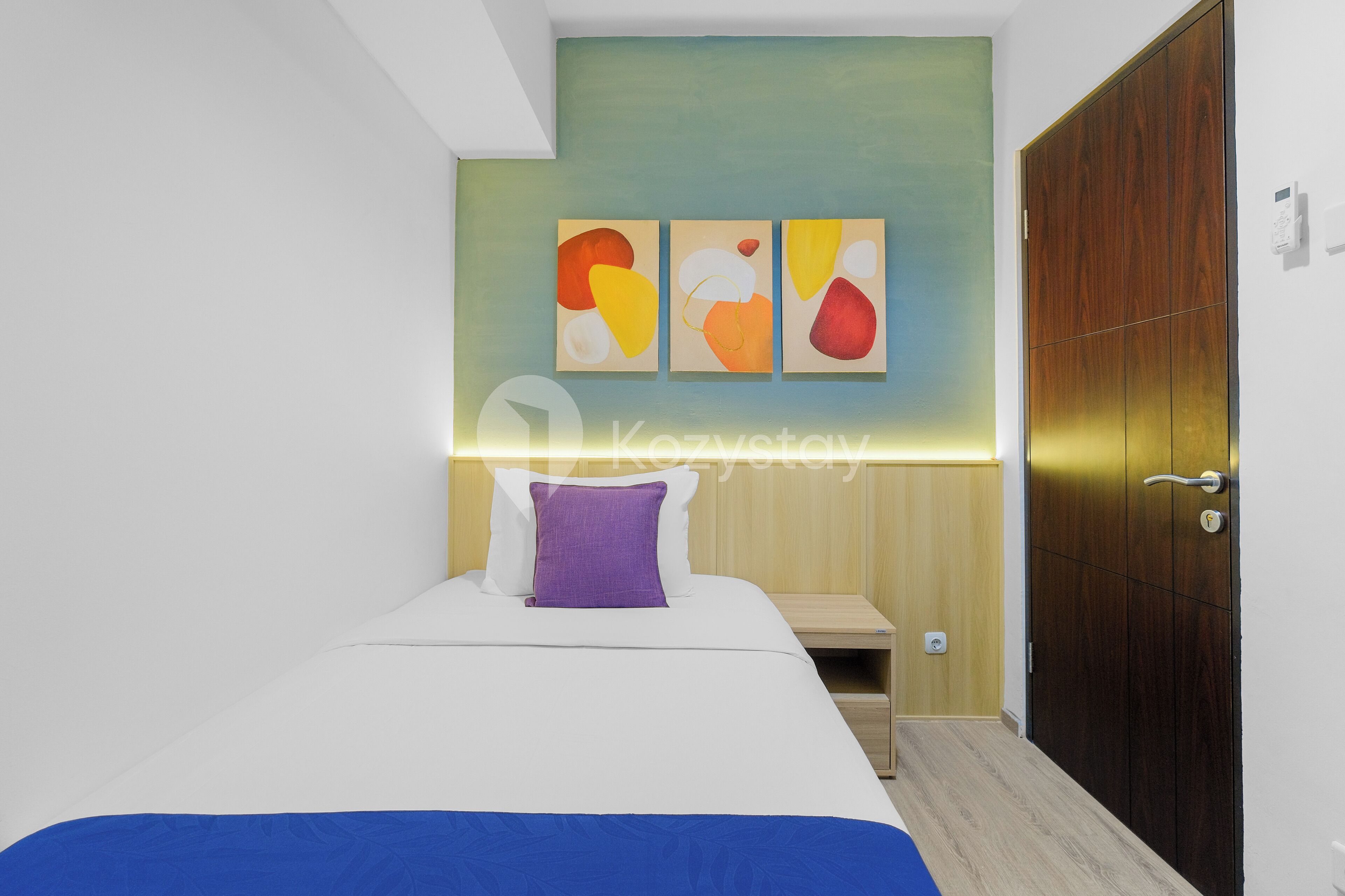 A modern hotel room with a bed, a nightstand, a wardrobe, and abstract wall art.