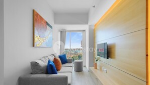 Una sala de estar moderna con un sofá gris, cojines coloridos, un televisor de pantalla plana y un amplio ventanal con vistas a la ciudad.