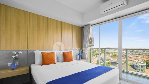 Una habitación de hotel moderna con una cama grande, cabecera de madera, mesita de noche con lámpara y vistas a la ciudad a través de amplios ventanales.