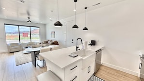 A modern kitchen with white cabinetry, a central island, pendant lighting, and a view of the outdoors through large windows. - Kanab Desert Oasis - Pool, Hot Tub + Pickleball (Kanab)