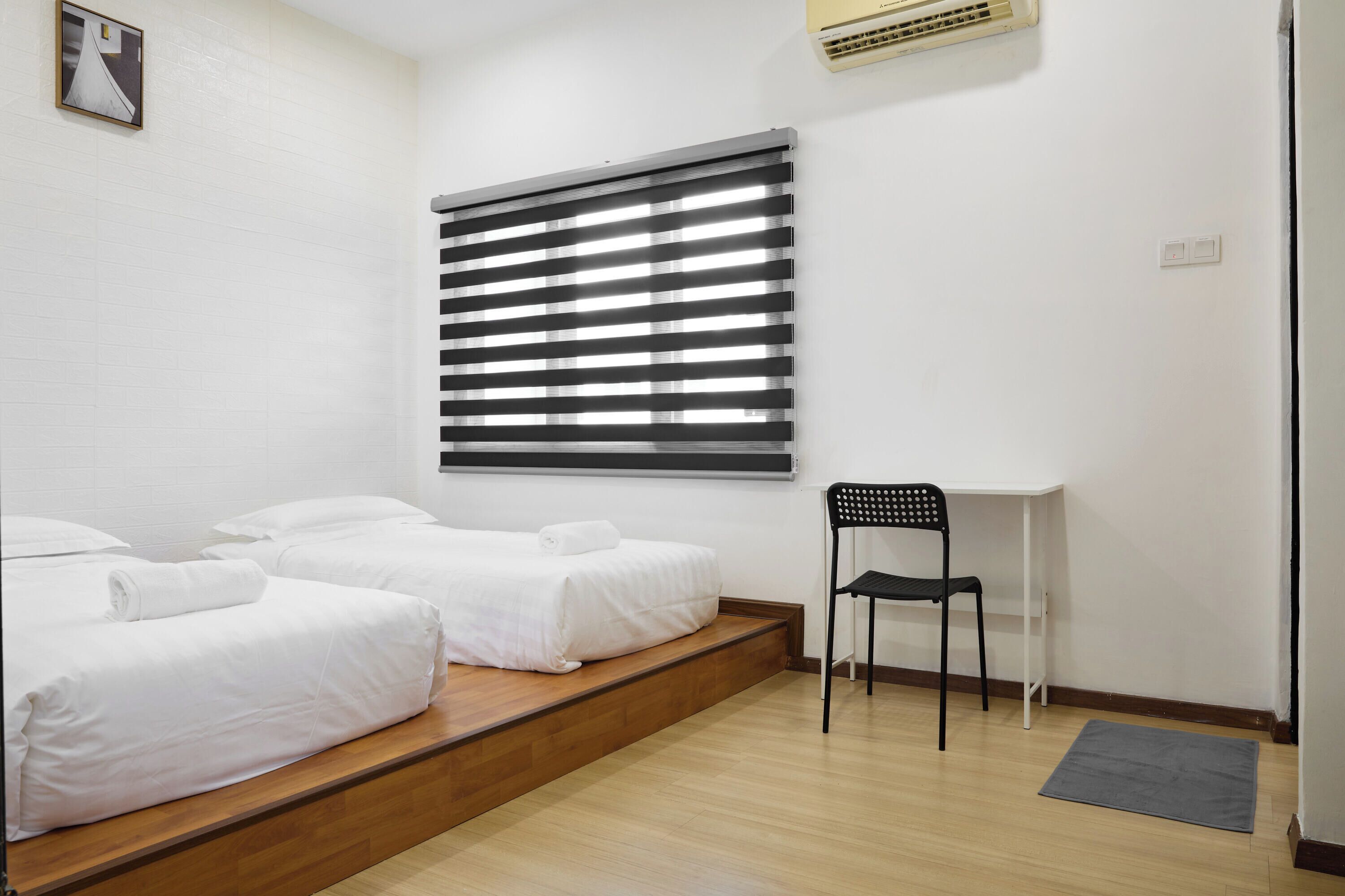 A room with two single beds, a desk, a chair, and a window with blinds.