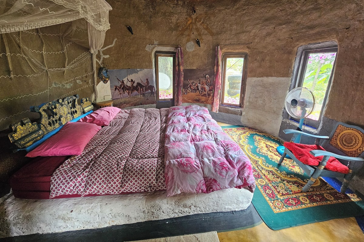 Habitació rústica amb un llit gran, una cadira, un ventilador i un mural a la paret.