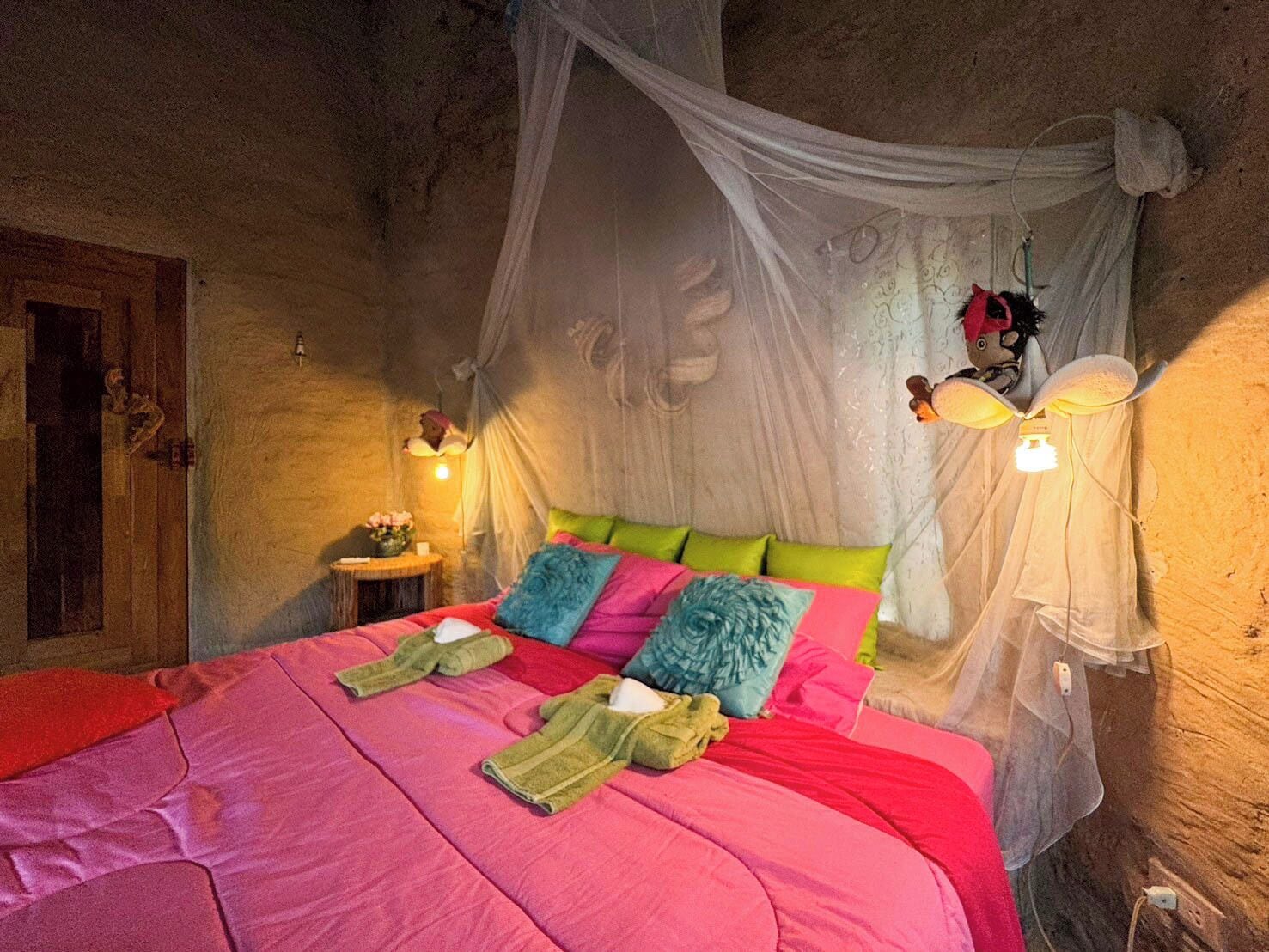 A room with a bed covered in a pink blanket, green and blue pillows, and a canopy. There are two bedside lamps and a small table with a lamp.