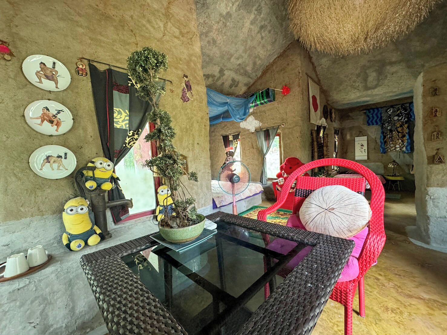 A room with a glass table, wicker chairs, a small potted plant, and various wall decorations including plates and figurines.