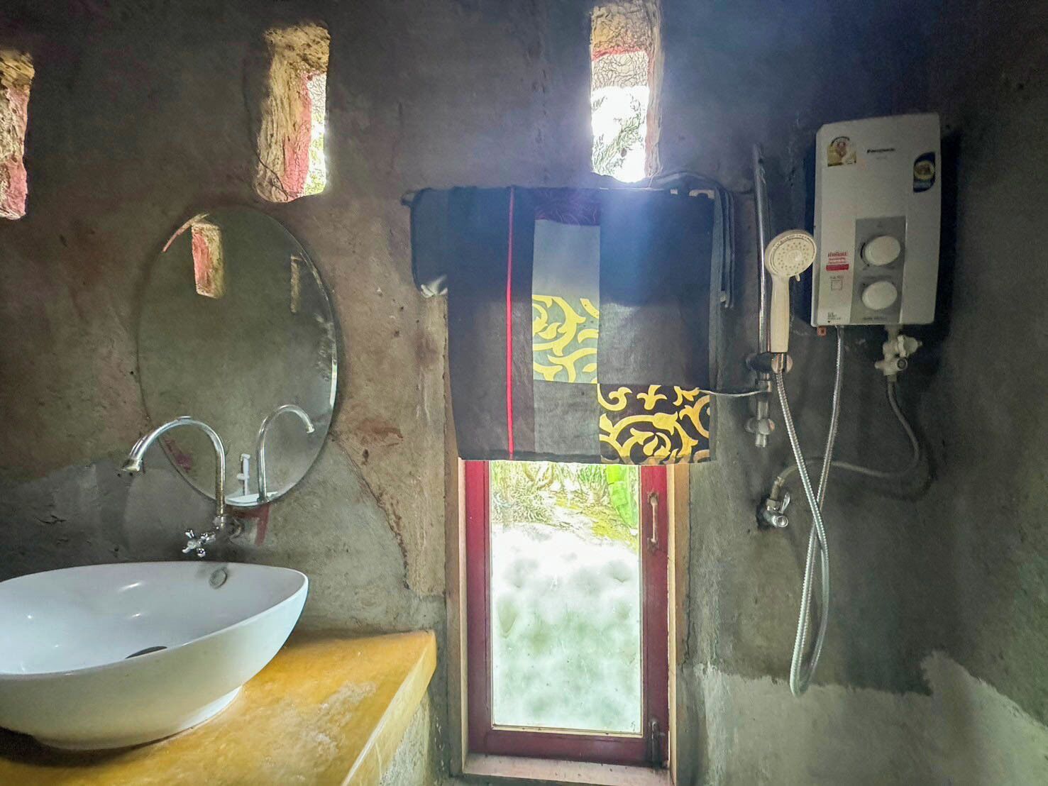 A bathroom with a white sink, a mirror, a shower, and a window with patterned curtains.