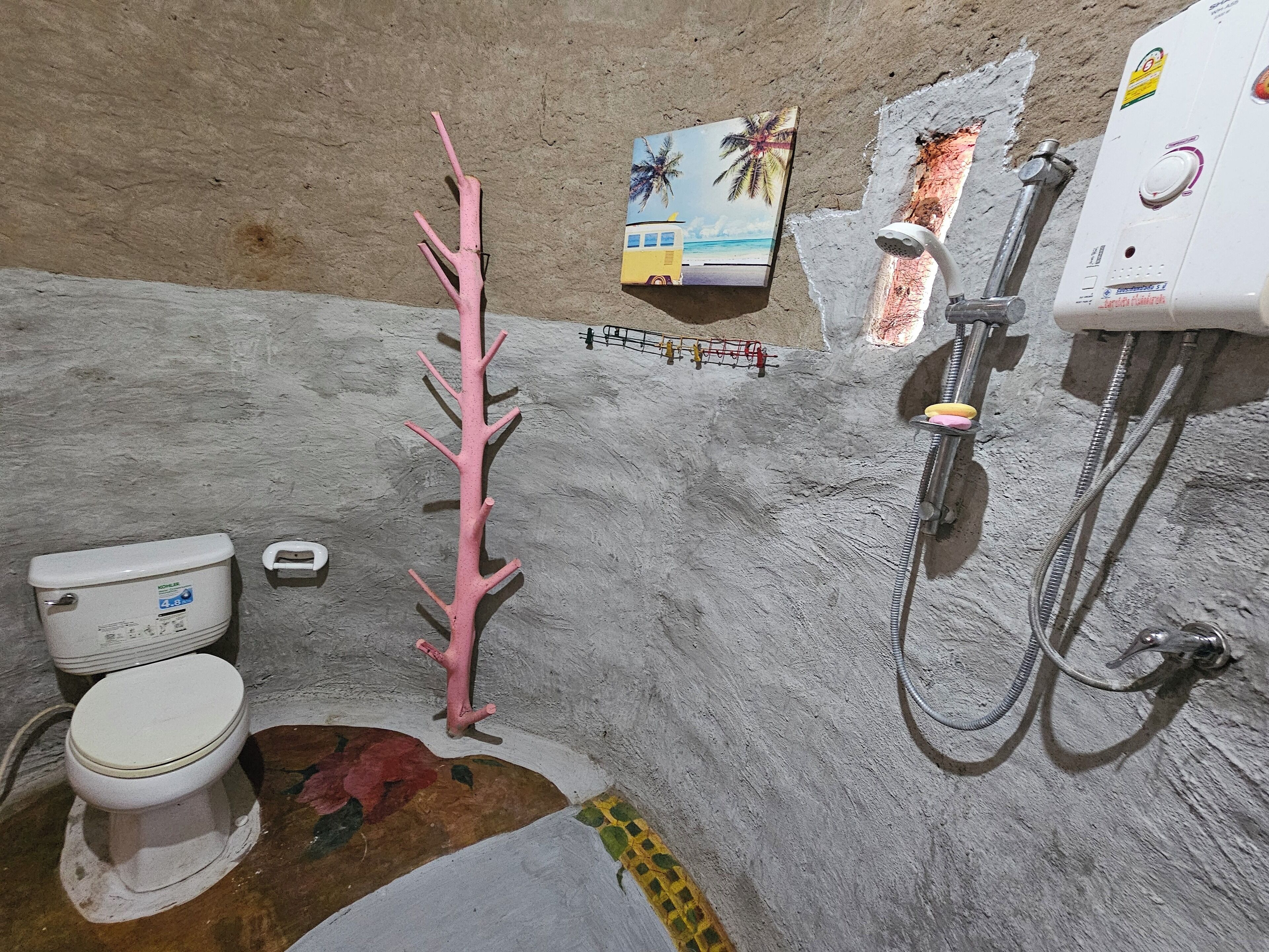 Et badeværelse med toilet, et pink dekorativt træ, bruser og en lille hylde med et billede.