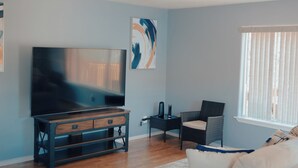 Ein Wohnzimmer mit einem Flachbildfernseher, einem weißen Sofa, einem Korbstuhl und einem großen abstrakten Gemälde an der Wand.