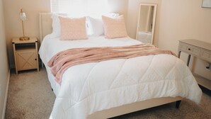 Ein ordentlich bezogenes Bett mit weißer Decke, zwei rosafarbenen Kissen und einer rosafarbenen Tagesdecke. Ein Nachttisch mit Lampe und einem kleinen Holzschrank sind vorhanden. An der Wand über dem Bett ist ein Spiegel angebracht.