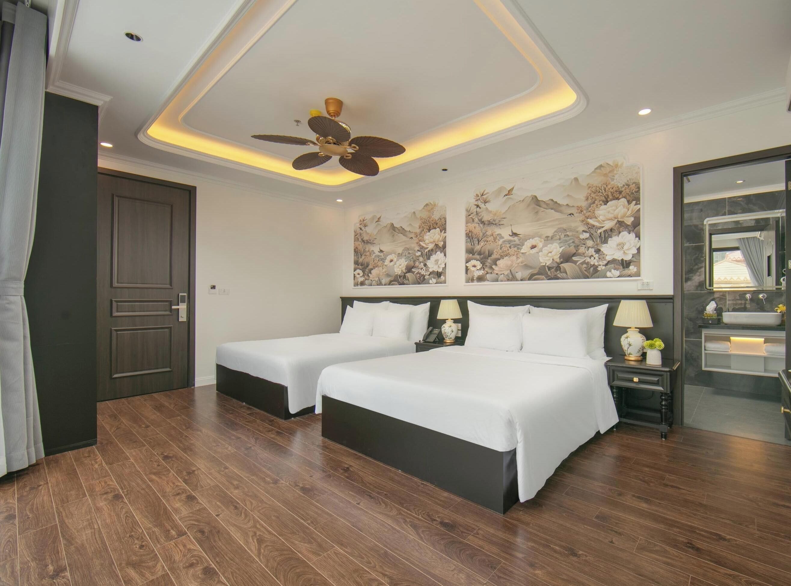 A hotel room with two beds, a ceiling fan, artwork on the walls, a nightstand with a lamp, and a bathroom with a mirror and sink.