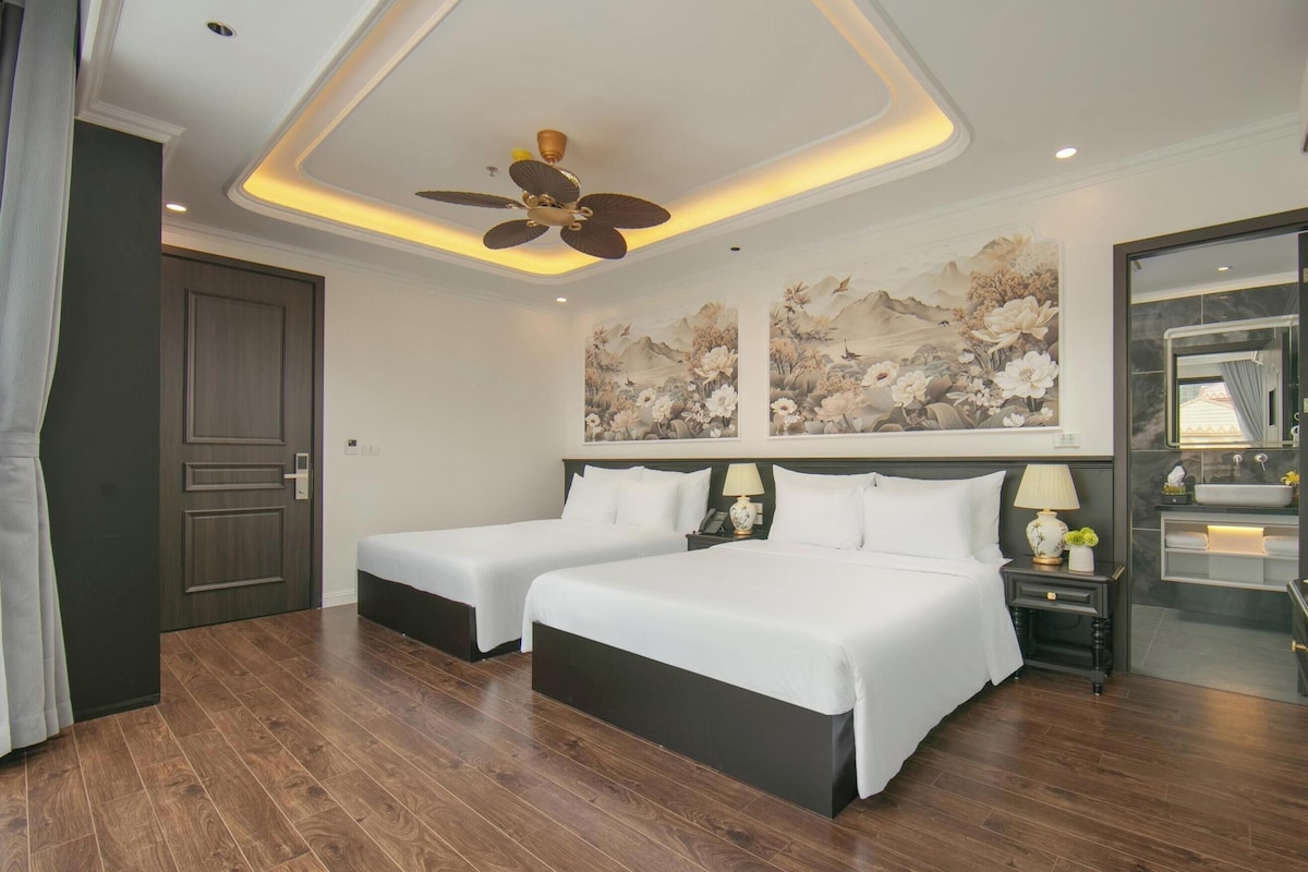 A hotel room with two beds, a ceiling fan, artwork on the walls, a nightstand with a lamp, and a bathroom with a mirror and sink.