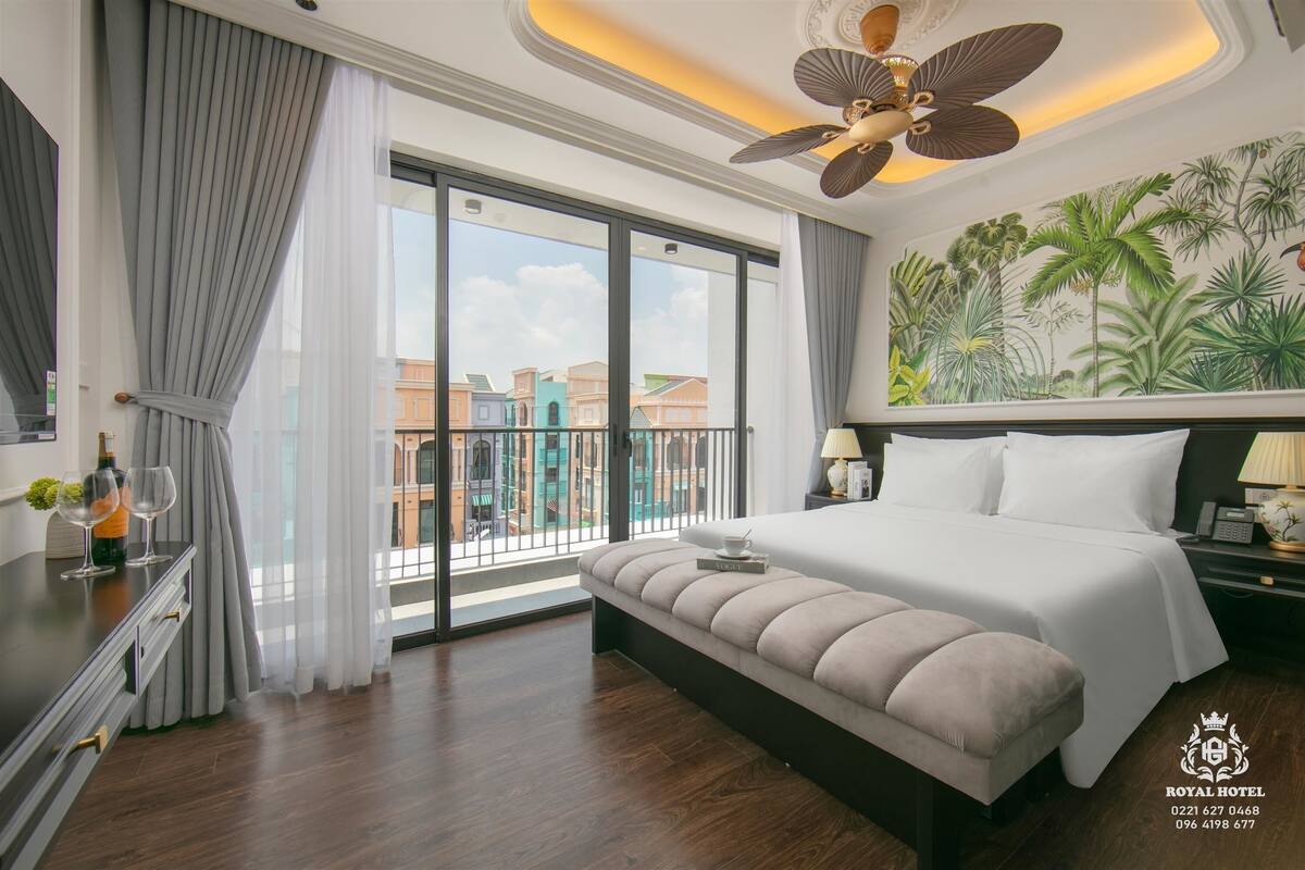 Habitación de hotel con una cama grande, un banco, un ventilador de techo, vistas a edificios a través de un ventanal y un mural tropical en la pared.