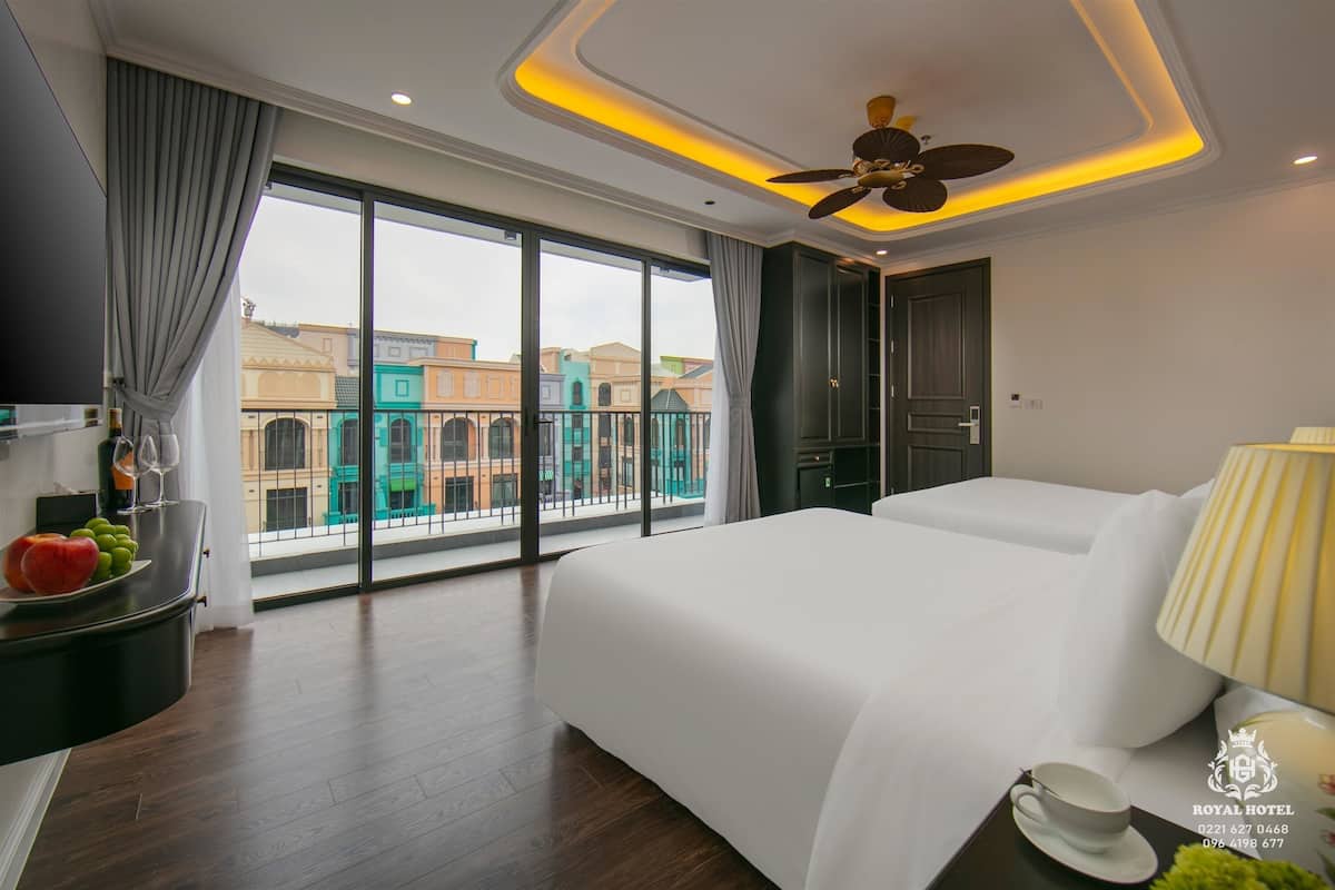 Una habitación de hotel con una cama grande, una mesita de noche con una taza y un plato, un ventilador de techo y vistas a edificios a través de la ventana.
