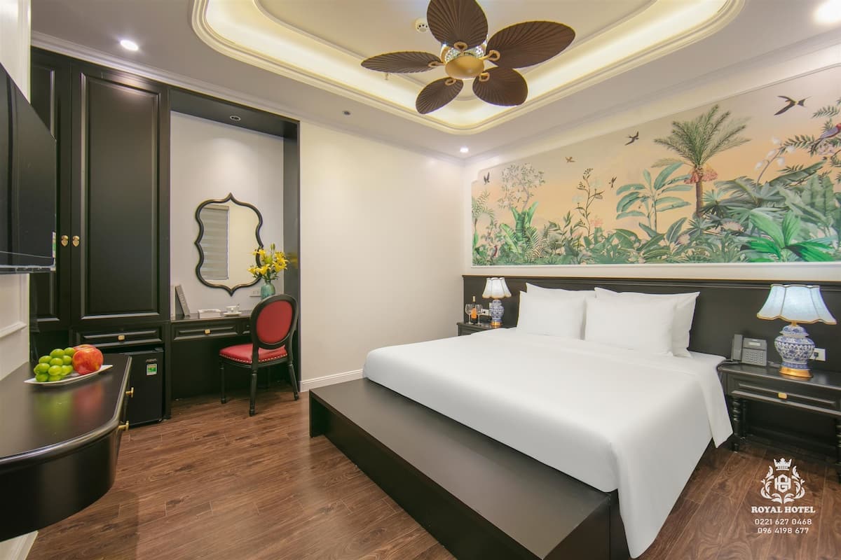 Habitación de hotel con una cama, un escritorio con una silla, una mesita con fruta, un espejo y un mural en la pared.