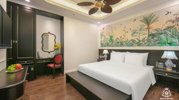 A hotel room with a bed, a desk with a chair, a small table with fruit, a mirror, and a wall mural.