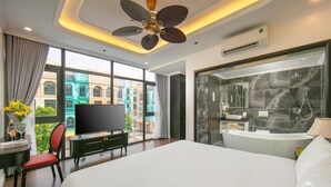 A modern hotel room with a large bed, a television, a desk with a chair, a ceiling fan, and a large window offering a city view. - Royal Hotel - Vinhomes Ocean Park 3 (Nghia Tru)