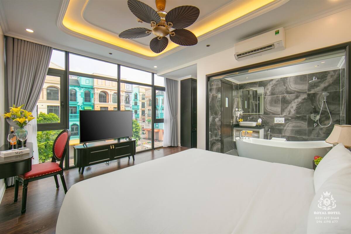 Una habitación de hotel moderna con una cama grande, televisor, escritorio con silla, ventilador de techo y una ventana amplia con vistas a la ciudad.
