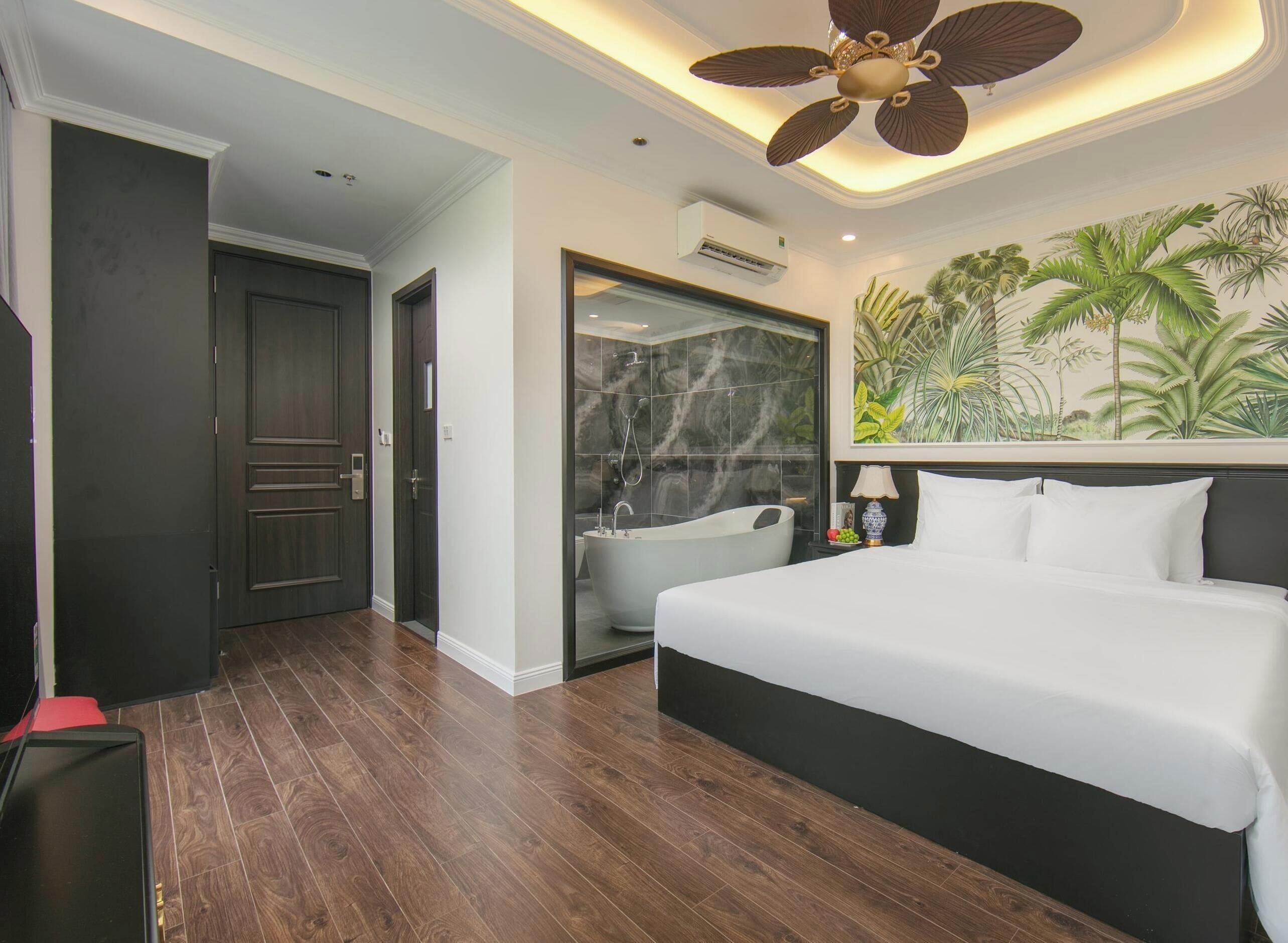 A hotel room with a large bed, a ceiling fan, a bathtub, and a mural of tropical plants.