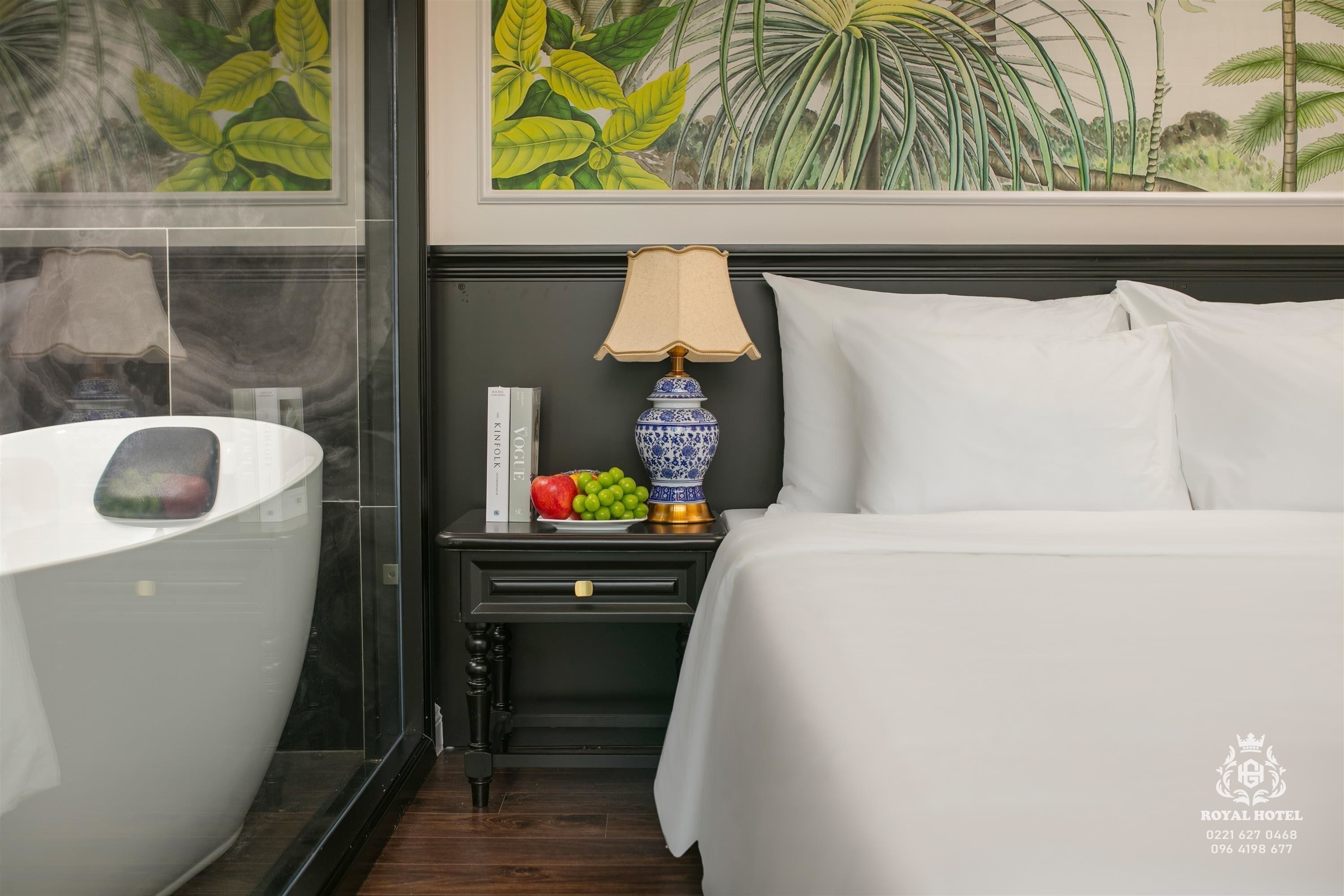 A hotel room with a bathtub, a bedside table with a lamp and fruit, a bed with white linens, and a tropical-themed wall mural.