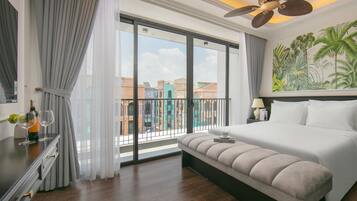 Habitación de hotel con una cama grande, un banco, un ventilador de techo, vistas a edificios a través de un ventanal y un mural tropical en la pared.