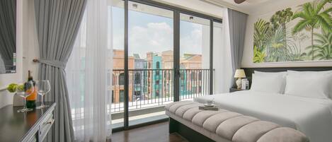 A hotel room with a large bed, a bench, a ceiling fan, a view of buildings through a glass door, and a tropical mural on the wall.