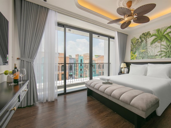 A hotel room with a large bed, a bench, a ceiling fan, a view of buildings through a glass door, and a tropical mural on the wall. - Royal Hotel - Vinhomes Ocean Park 3 (Nghia Tru)