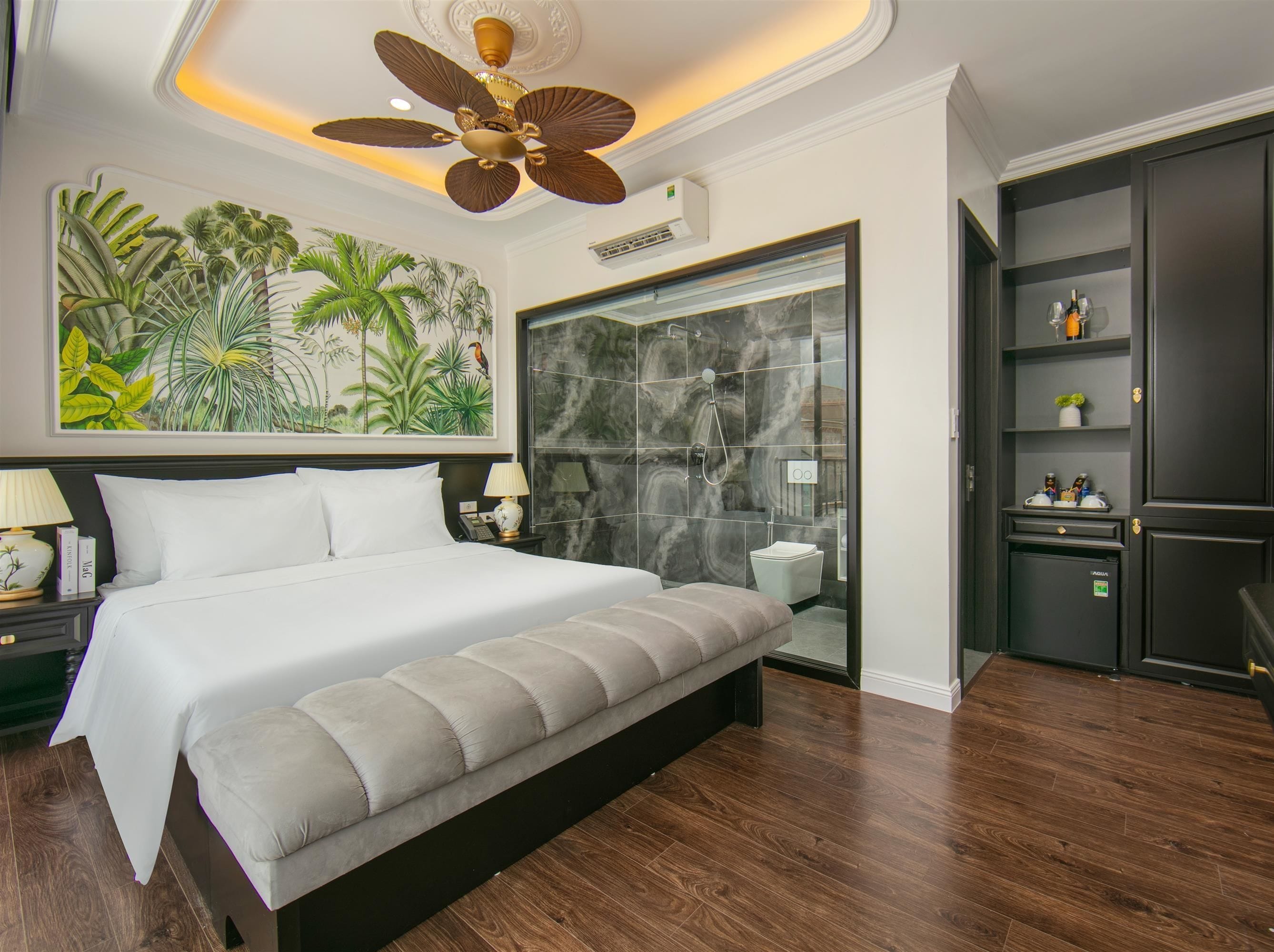 A modern hotel room with a large bed, a ceiling fan, a tropical mural, a bathroom with a glass door, and a minibar.