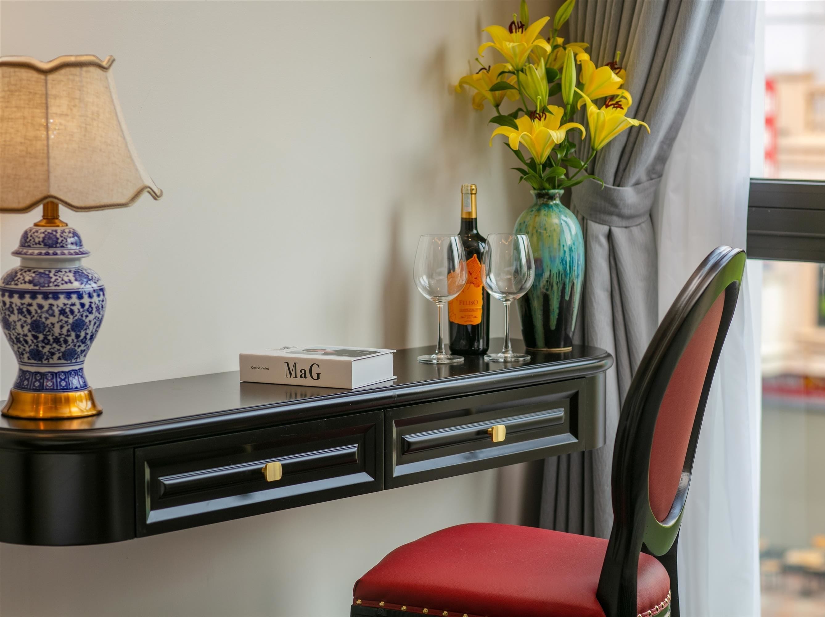 A hotel room with a desk, a red chair, a lamp, a vase with yellow flowers, and a bottle of champagne.