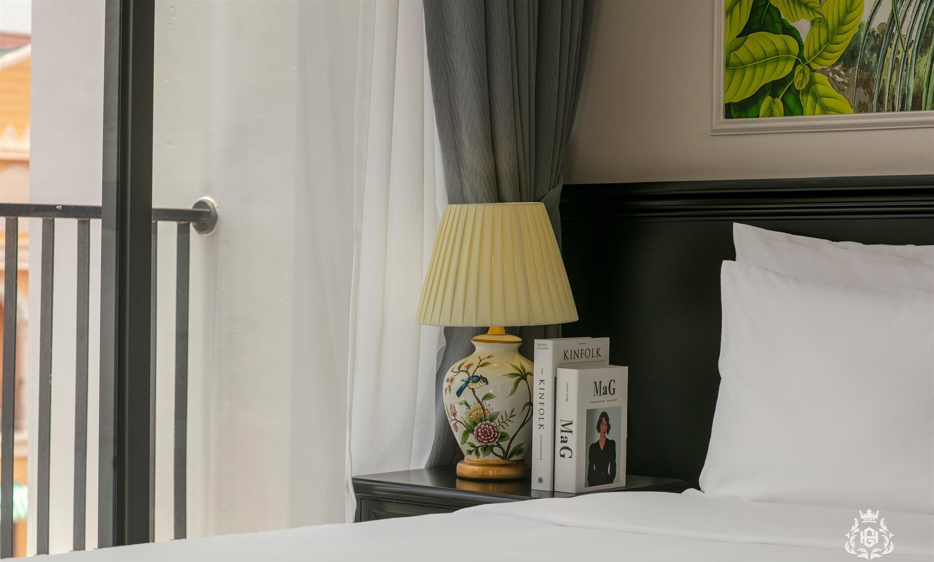 A neatly made bed with white linens, a black headboard, a decorative lamp with floral patterns, and two books titled 'KINFOLK' and 'MaG'.