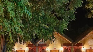 Un bâtiment traditionnel doté de nombreuses portes et fenêtres en bois, entouré d’une végétation luxuriante et d’un petit étang.