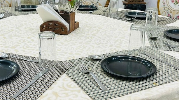 Une table de salle à manger ronde, recouverte d’une nappe blanche, avec des assiettes noires et des couverts en argent. Des assiettes décoratives ornées de motifs floraux sont disposées sur le bord de la table.