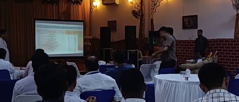 A conference room with attendees seated at round tables, a presentation screen, and speakers at the front.