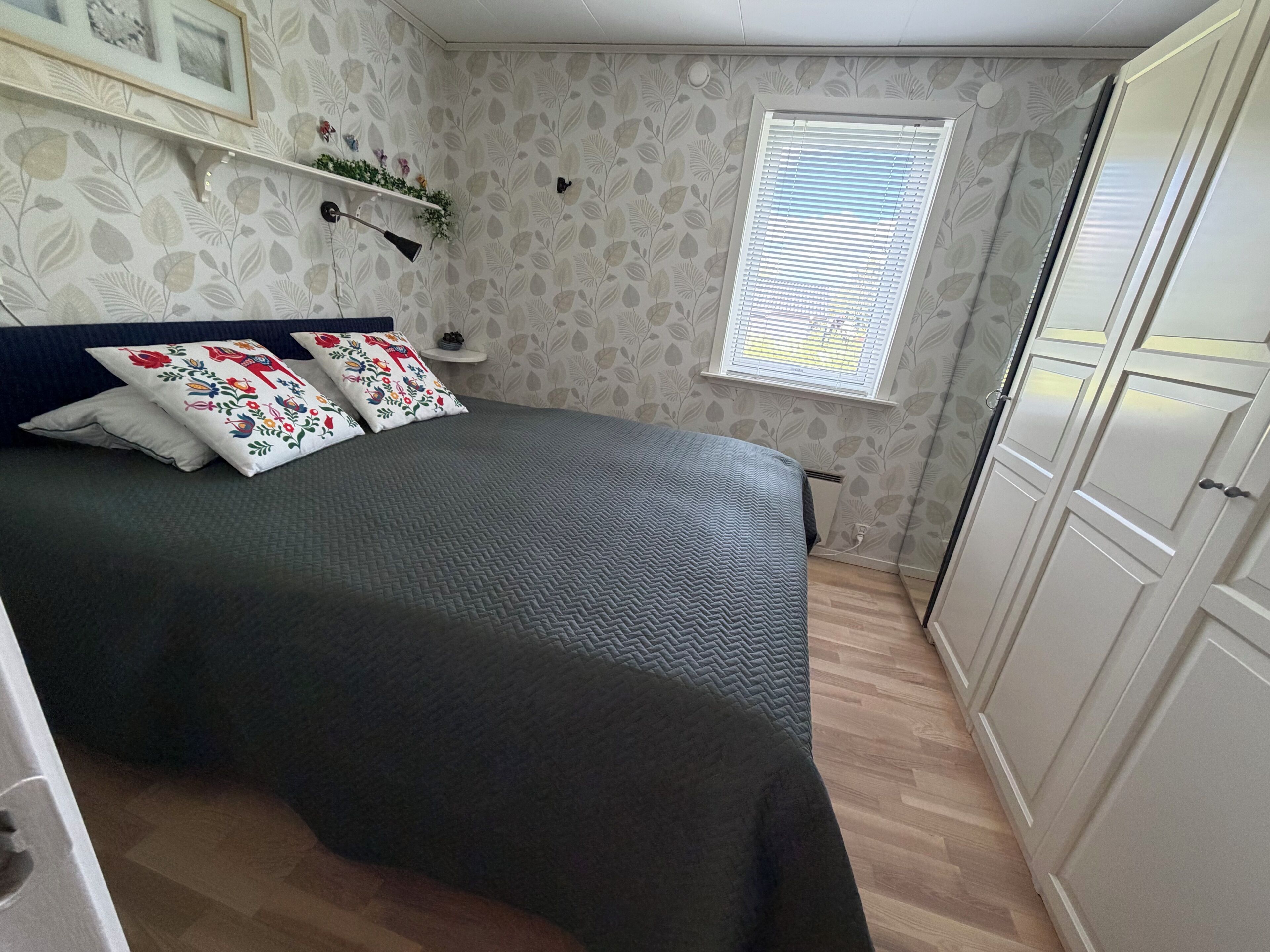 A bedroom with a bed, a window, a wardrobe, and a shelf with decorative items.