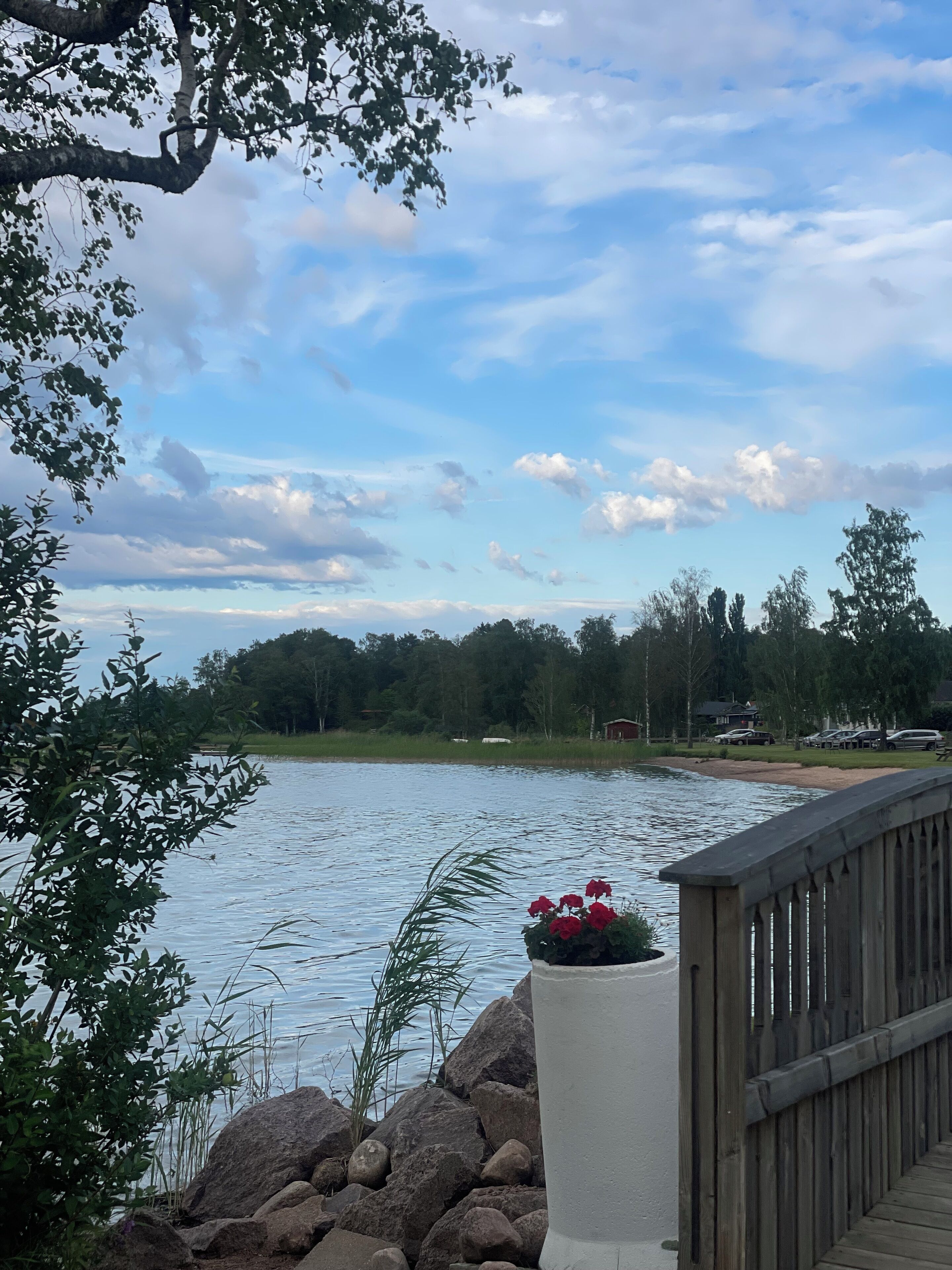 Vistes a un llac amb un pont de fusta, una jardineria de pedra amb flors vermelles i un fons d'arbres i una casa allunyada.