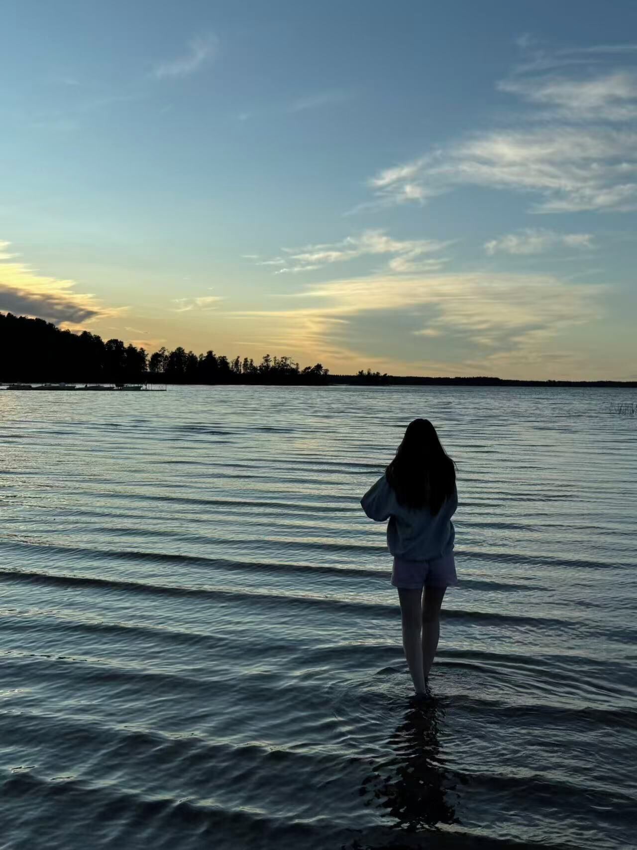 Une personne se tient dans une eau peu profonde au coucher du soleil, avec une côte et des arbres visibles au loin.
