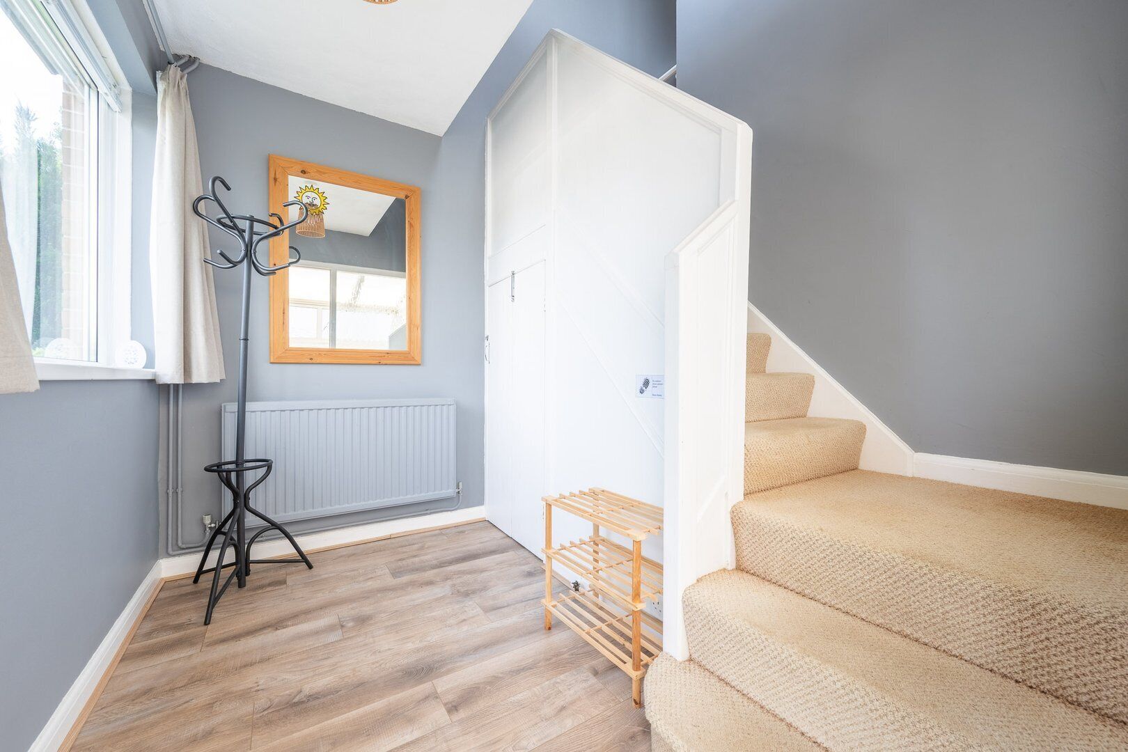 Habitación con piso de madera, un radiador, perchero, espejo y una escalera con alfombra color crema.