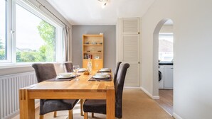 A dining area with a wooden table set for four, flanked by two chairs, a bookshelf, and a door leading to a kitchen. - Charming 2-Bed Detached Home in Radyr (Cardiff)