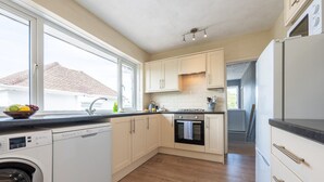 A compact kitchen with a dishwasher, oven, and a window with a view of a house. - Charming 2-Bed Detached Home in Radyr (Cardiff)