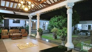 Un espacio al aire libre cubierto con un banco de madera y plantas en macetas, suelo de baldosas y una estructura de columnas blancas.