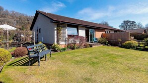 A single-story house with a sloped roof, a front porch, and a small garden area with a bench. - 3 bedroom accommodation in Pitlochry (Pitlochry)