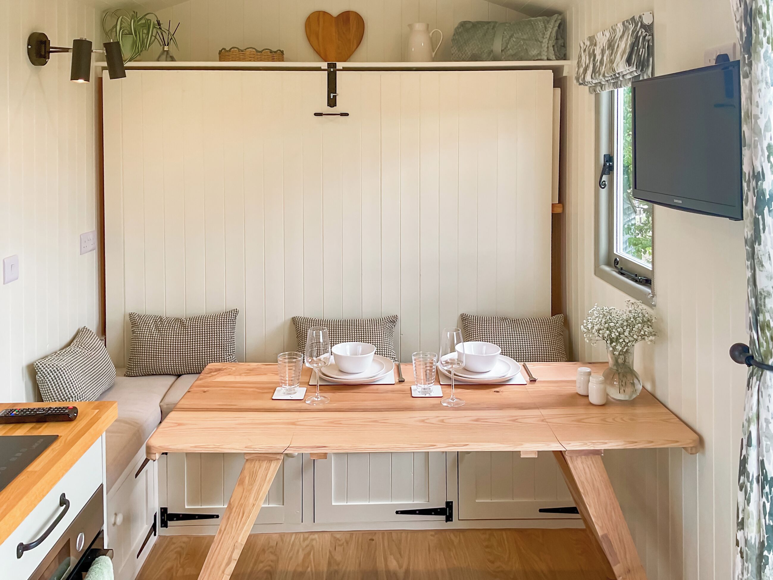 Un comedor con una mesa de madera dispuesta para dos personas, paredes blancas y un televisor montado en la pared.