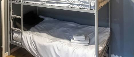 Un lit superposé avec une literie blanche, une petite étagère et une pile de vêtements pliés.