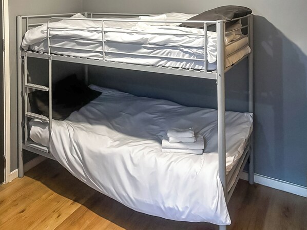 A bunk bed with white bedding, a small shelf, and a stack of folded clothes.