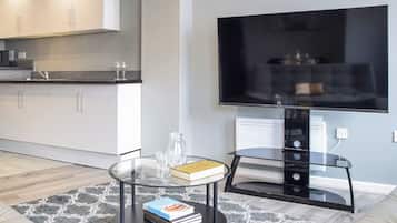 A modern living room with a glass coffee table, a flat-screen TV, a sofa, and a kitchen area in the background.