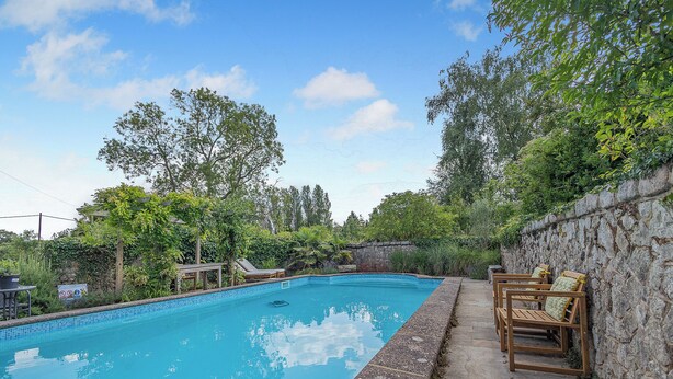 Piscina com parede de pedra e cadeiras de madeira, cercada por vegetação.