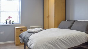 A bedroom with a bed, a wooden wardrobe, a bedside table, and a window with flowers. - Apartment 3 - UK50137 (Stoke-on-Trent)