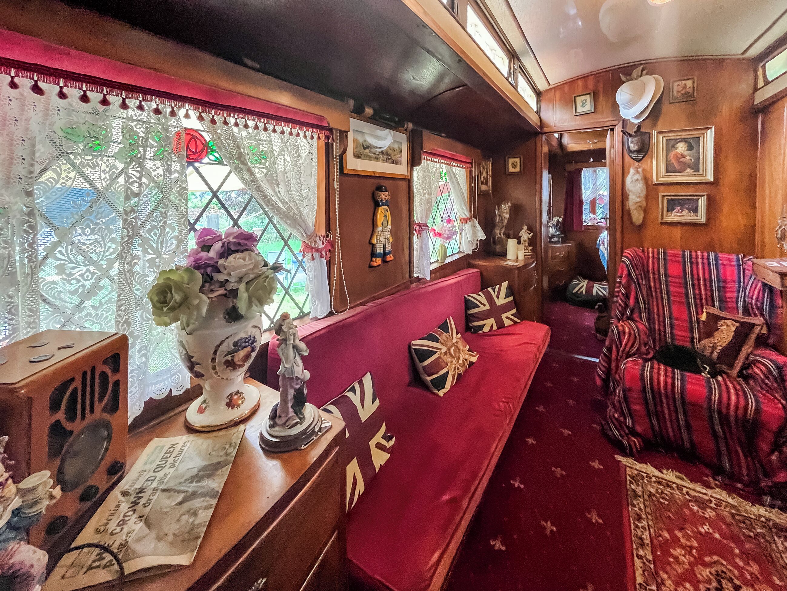 A train carriage interior with a red sofa, a table with a vase of flowers, and a window with lace curtains.