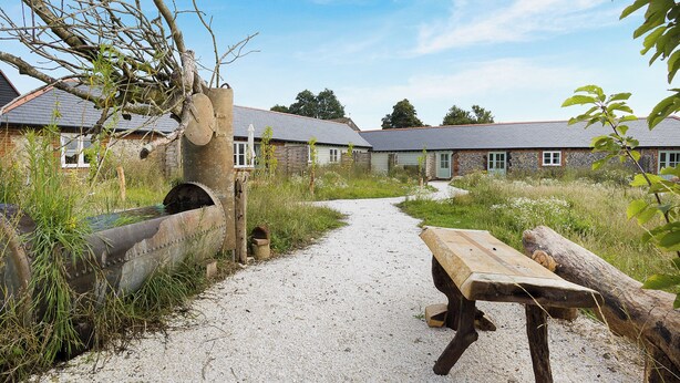 A gravel path leading to a rustic wooden bench and a stone structure, surrounded by overgrown vegetation and traditional buildings with sloped roofs. - 1 bedroom accommodation in Faversham (Faversham)
