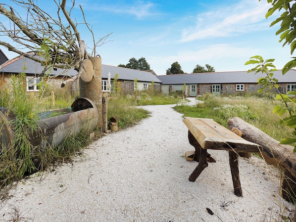 A gravel path leading to a rustic wooden bench and a stone structure, surrounded by overgrown vegetation and traditional buildings with sloped roofs.